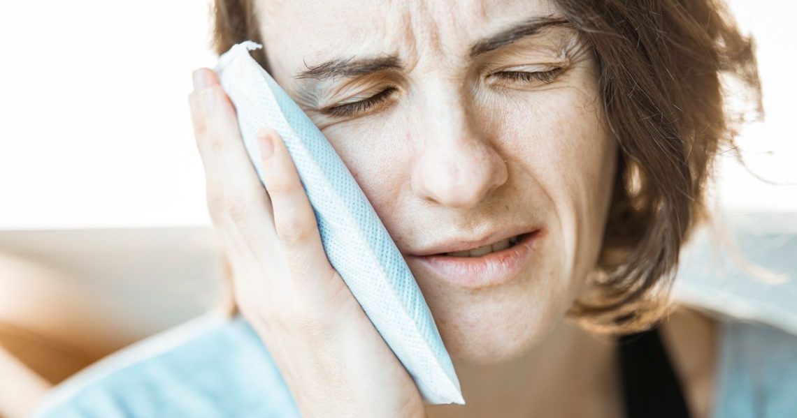 Dry Socket: Symptoms, Causes, and How to Prevent It 1 Woman wincing while holding a cold compress against her cheek